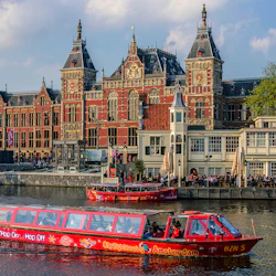 En röd sightseeingbåt navigerar en kanal med en stor, utsmyckad tegelbyggnad i bakgrunden och människor som går längs vattnet.