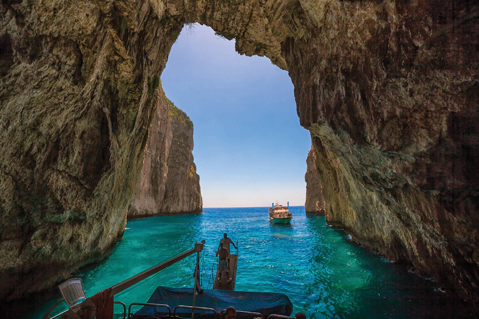 A boat approaches the entrance of a large sea cave, with turquoise water and clear blue sky visible through the opening.
