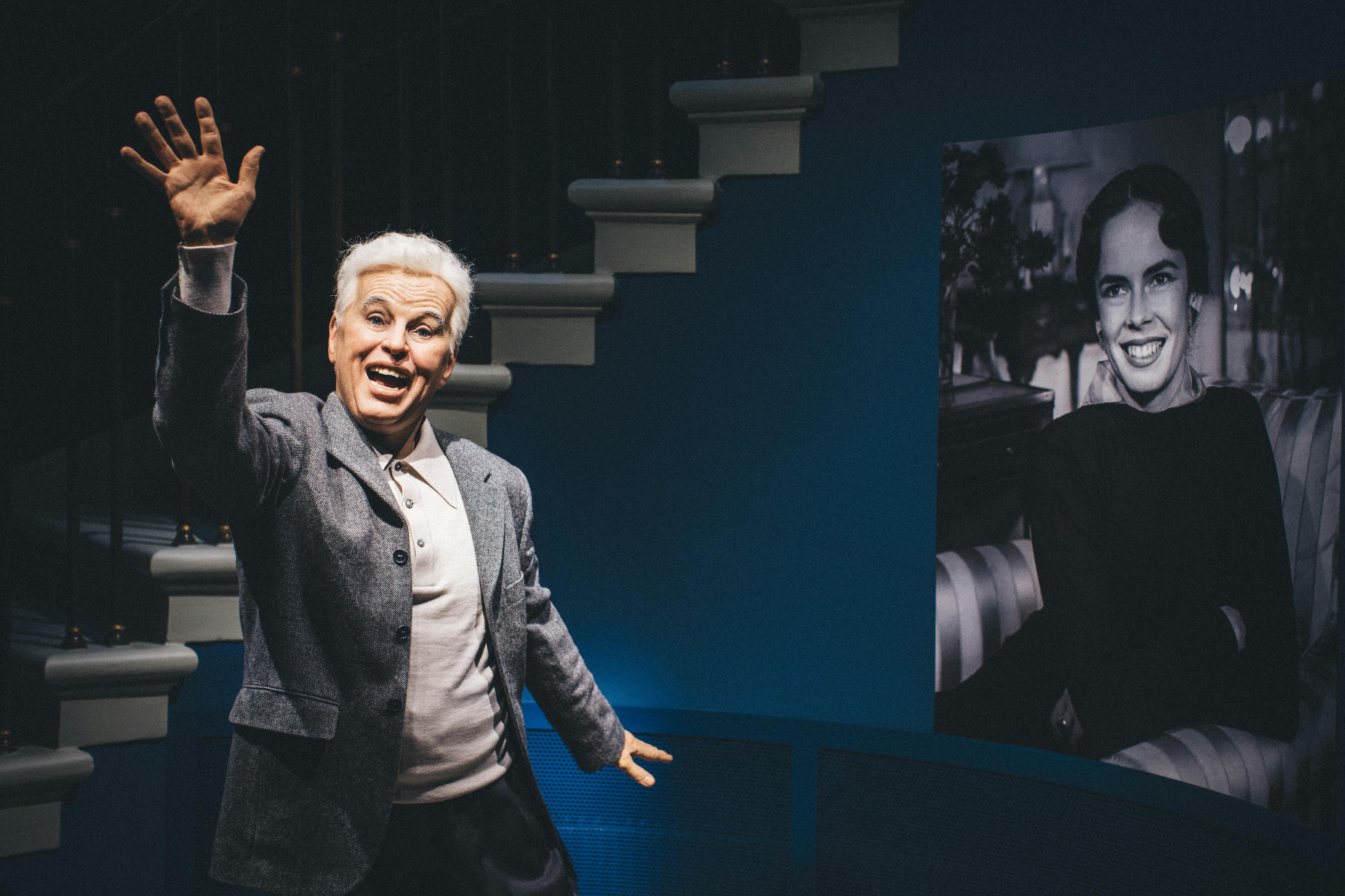 Před zakřiveným schodištěm stojí starší muž s bílými vlasy, šedým kabátem a světlou košilí, usmívá se a mává.