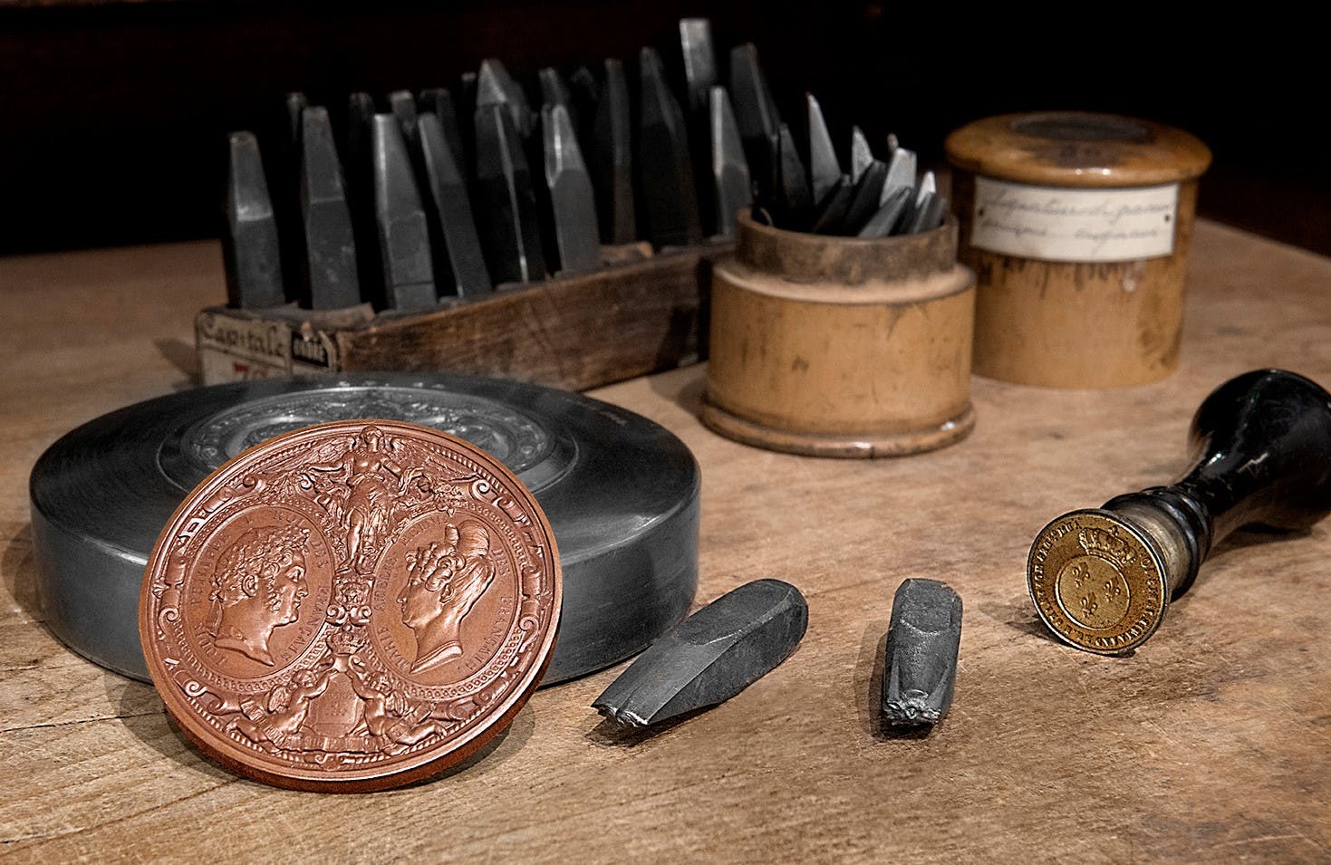 Κοντινό πλάνο των εργαλείων χάραξης, ένα μεγεθυμένο νόμισμα με δύο προφίλ προσώπου, ένα χρυσό νόμισμα και διάφορες σμίλες σε ένα ξύλινο τραπέζι.