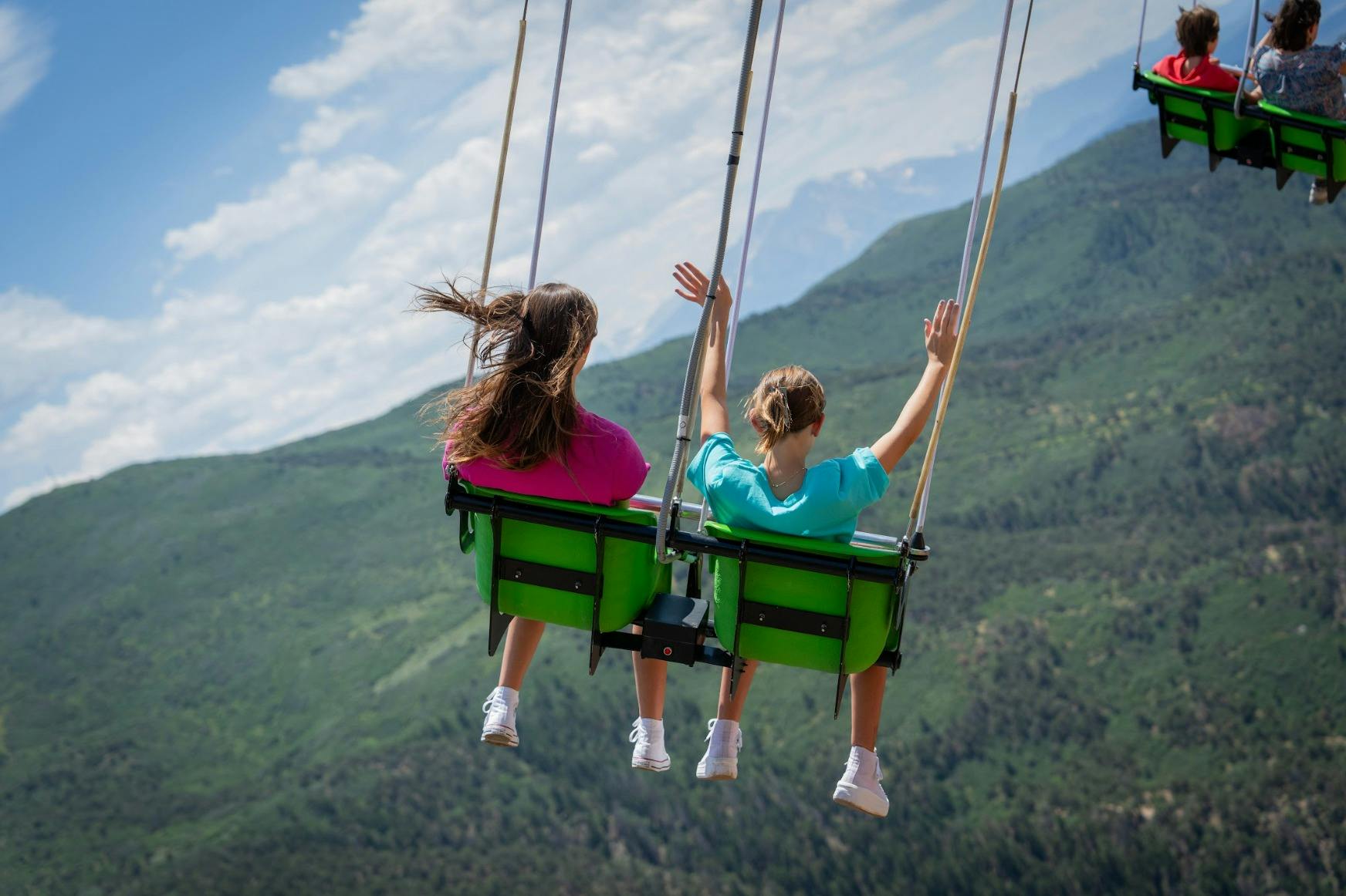 緑の山々と部分的に曇った空の風光明媚な景色を見下ろす、手を上げてブランコに乗っている2人。