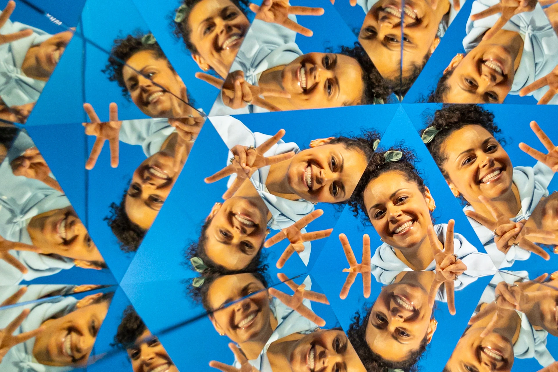 A kaleidoscopic image of a smiling person making peace signs, with repeated reflections creating a geometric pattern.