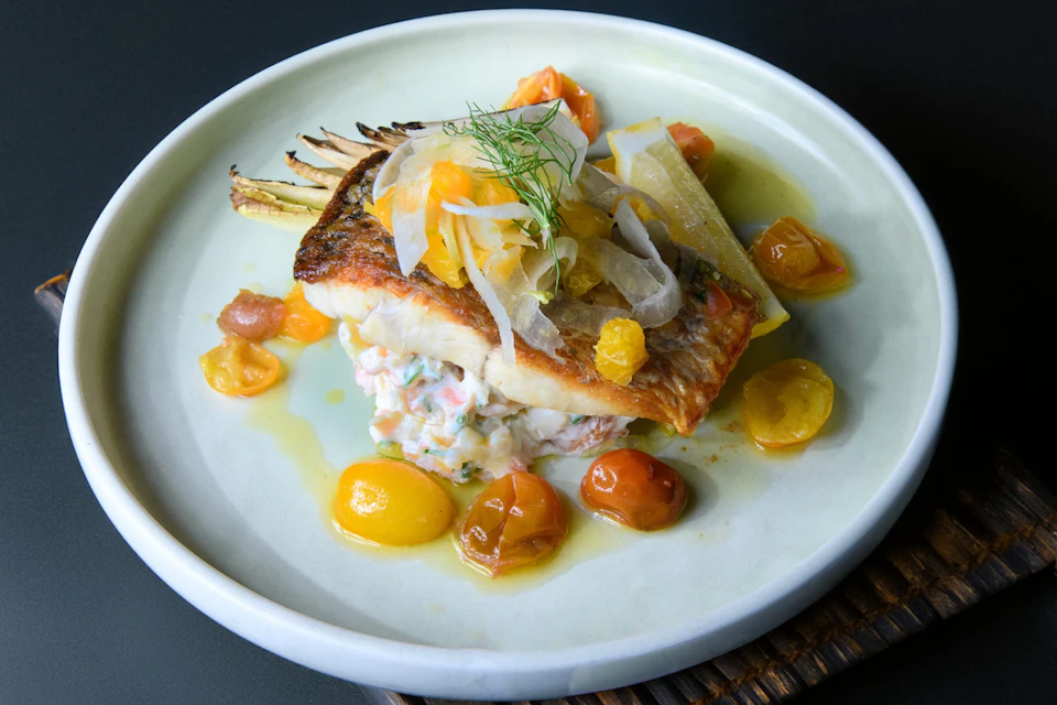 A plated dish featuring seared fish with toppings, cherry tomatoes, and a lemon wedge, served on a light-colored plate.