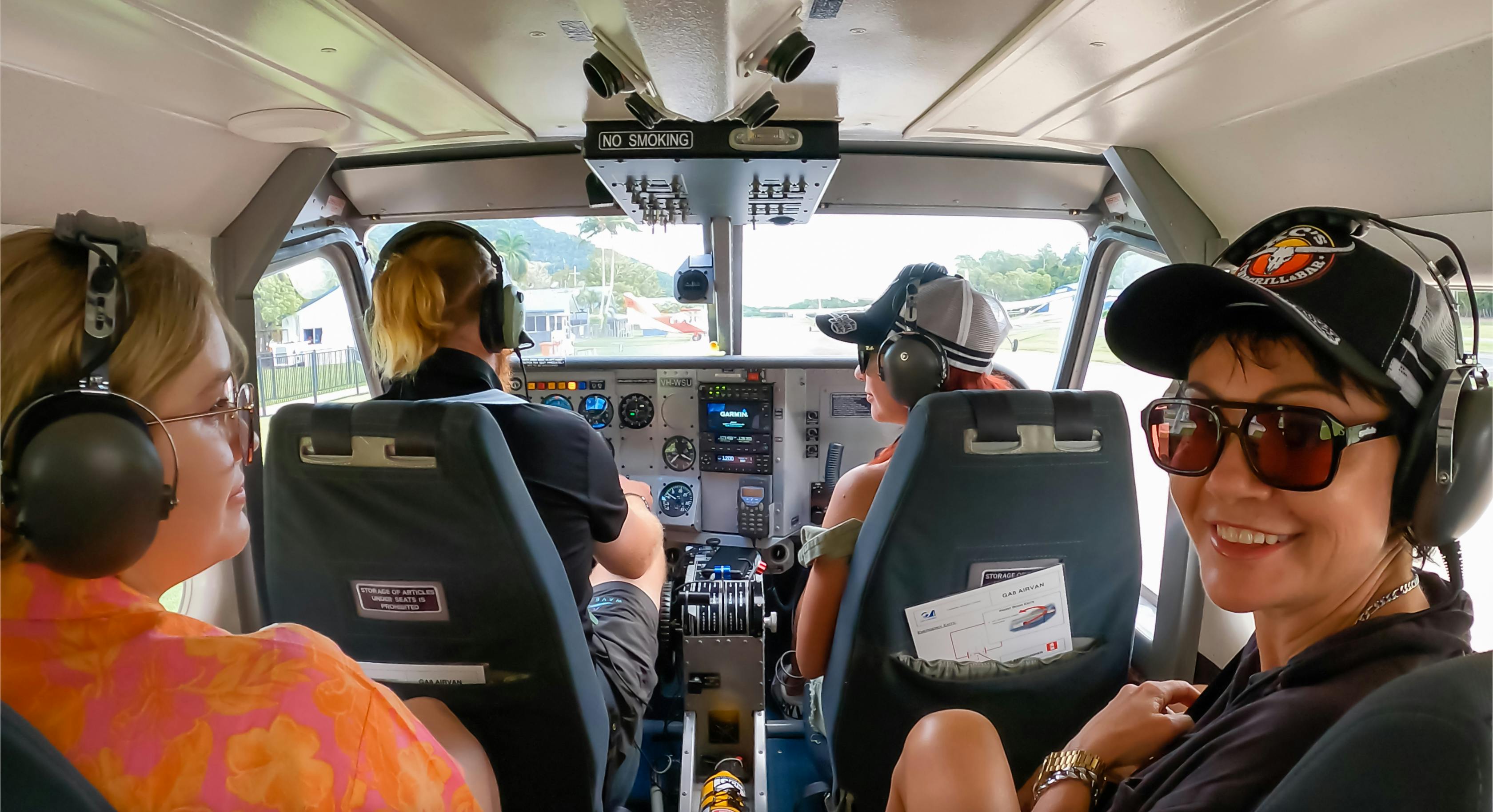 Two people in a small airplane cockpit. One is smiling at the camera wearing sunglasses and a cap; the other is piloting.