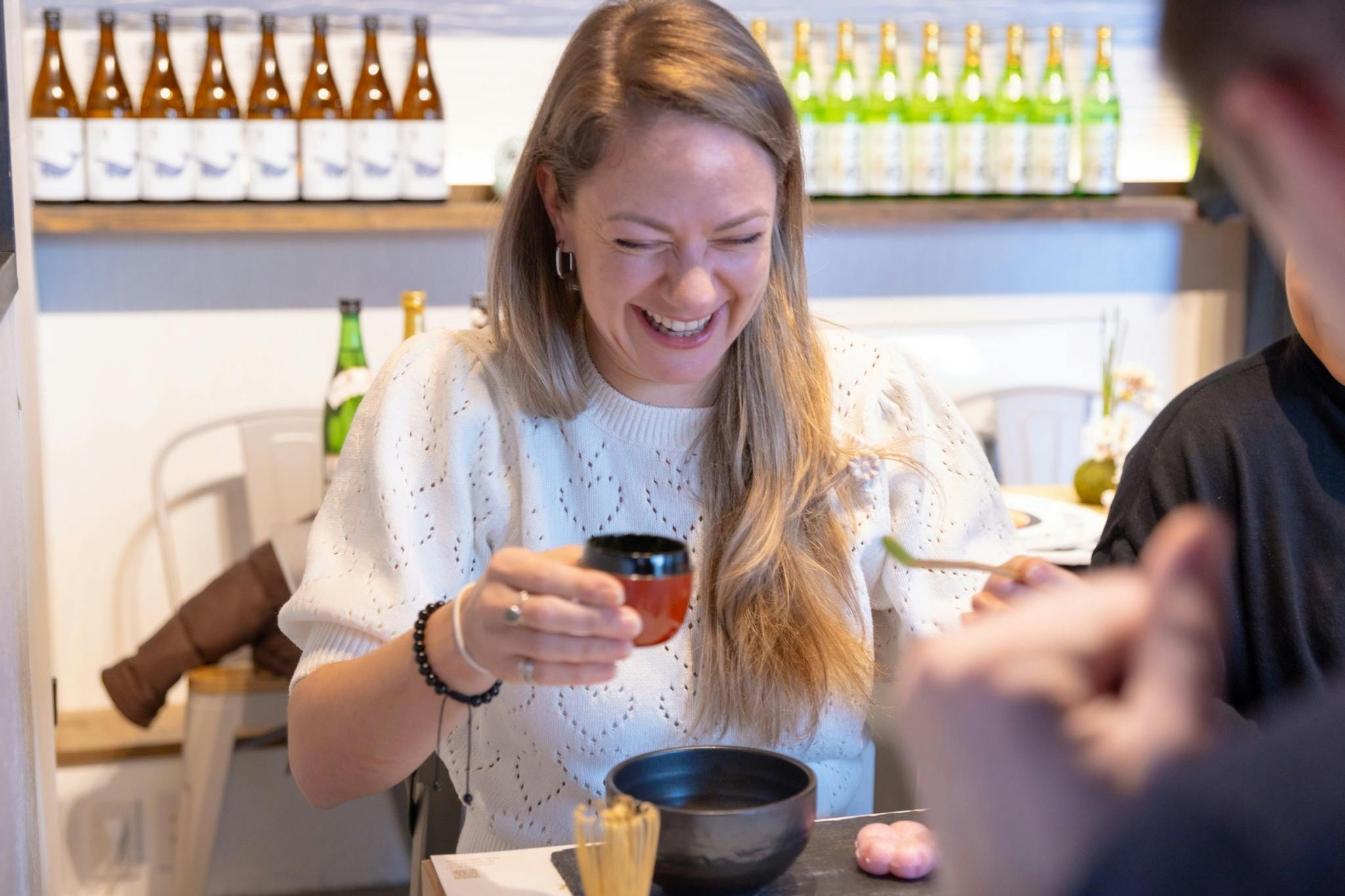 A smiling woman holds a small cup and sits at a table with bowls, food, and bottles displayed in the background.