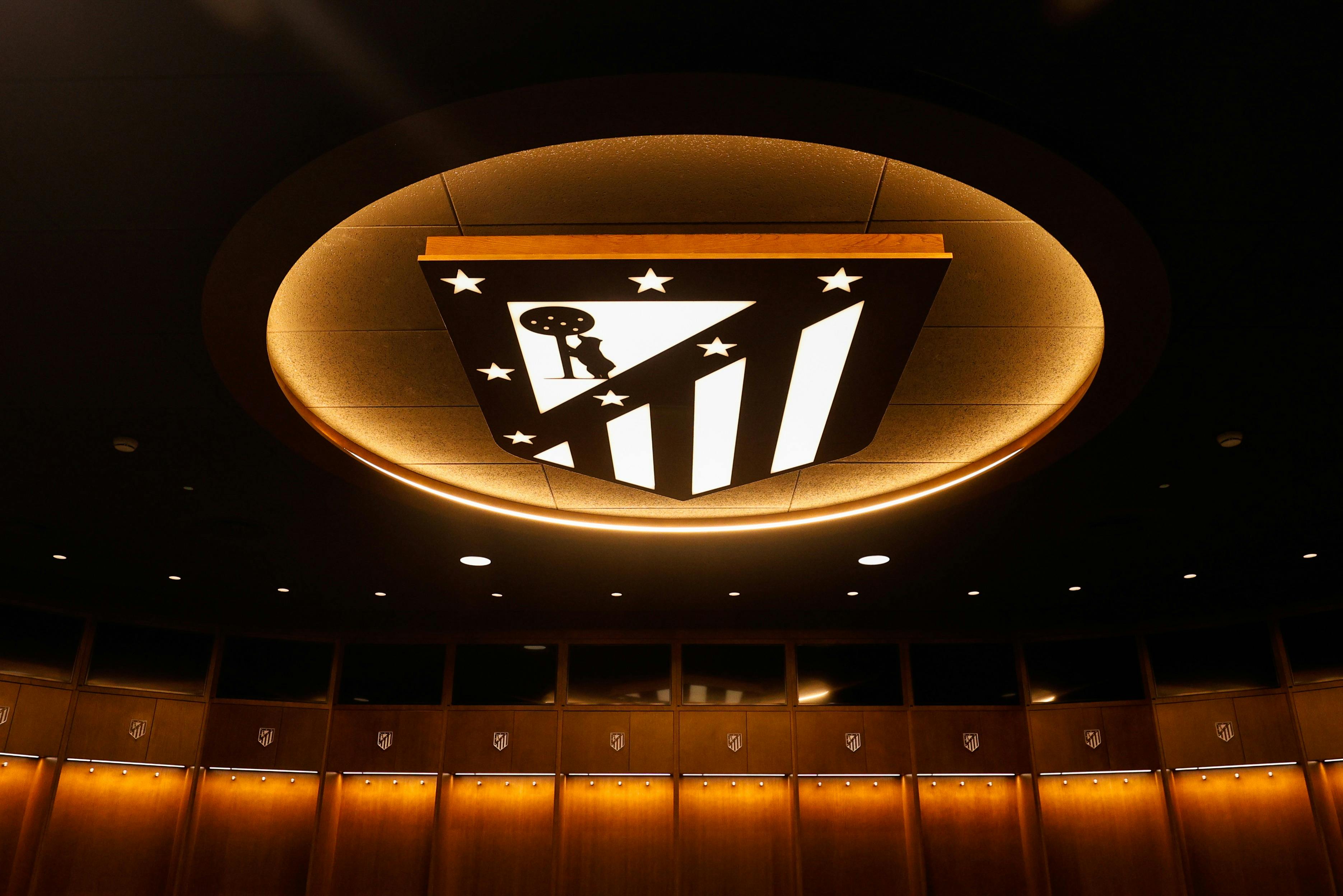 Een plafondlamp in de vorm van het logo van Atlético Madrid, boven een rij houten kastjes met de schildemblemen van de club.