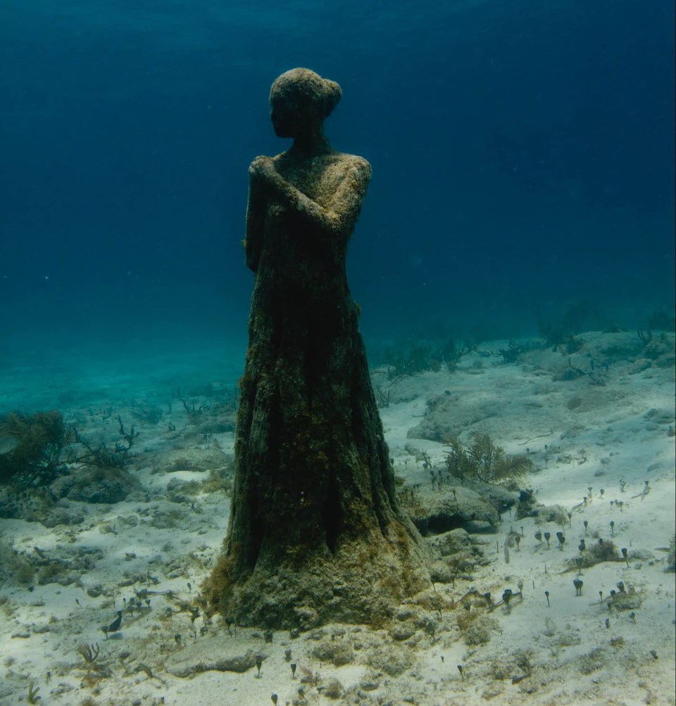 An underwater statue of a standing woman, encrusted with algae, on a sandy ocean floor with sparse coral and marine life.