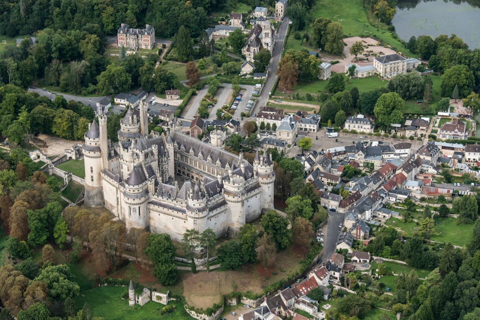 Aerial view of a large, medieval-style castle surrounded by trees, small buildings, and roads in a quaint village setting.