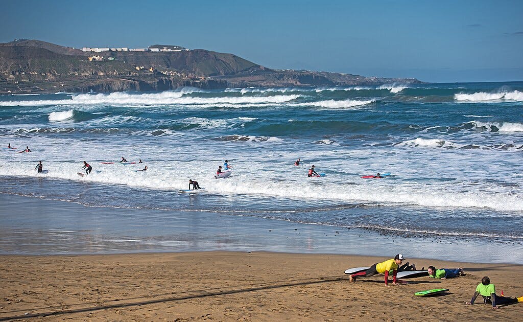 Люди занимаются серфингом и играют на песчаном пляже с волнами и далекими холмами под ясным голубым небом.