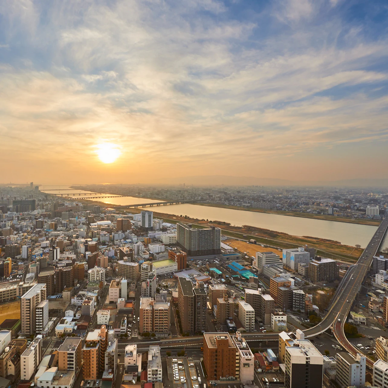 A cityscape with a river running through it, seen during sunset, featuring buildings, a bridge, and a dramatic sky.