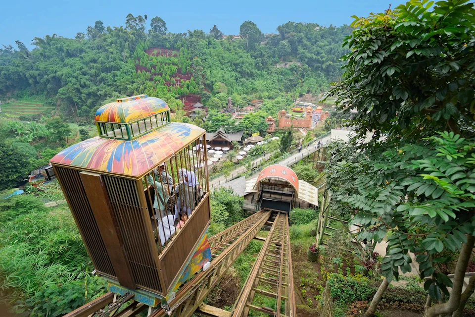 A colorful rail car transports people up a steep incline surrounded by lush greenery and a scenic village below.