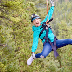 A person wearing a helmet and harness is ziplining through a forested area, smiling enthusiastically.