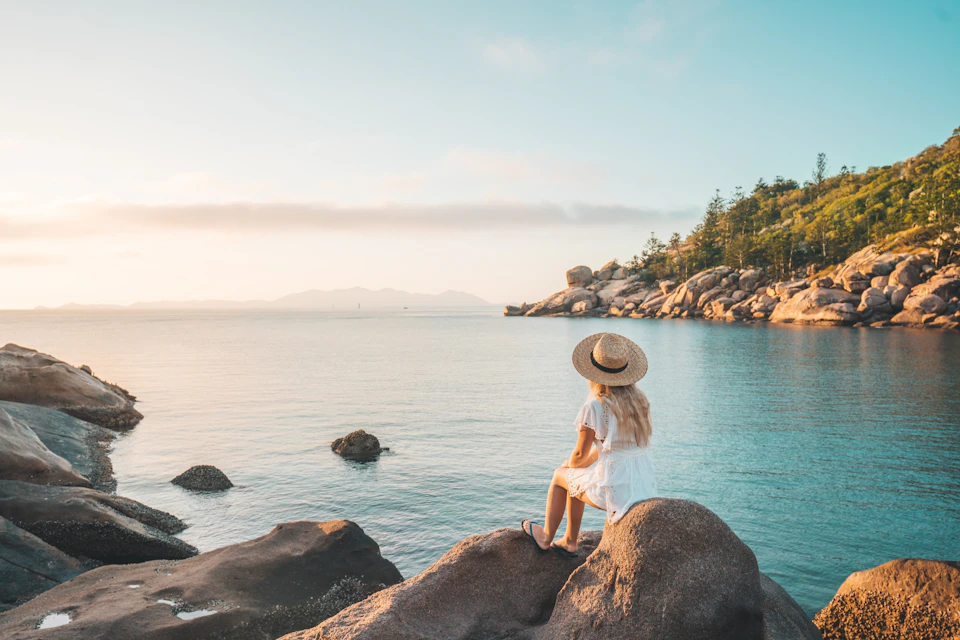 A person in a white dress and straw hat sits on a rock by calm water, with distant hills and trees under a clear sky.