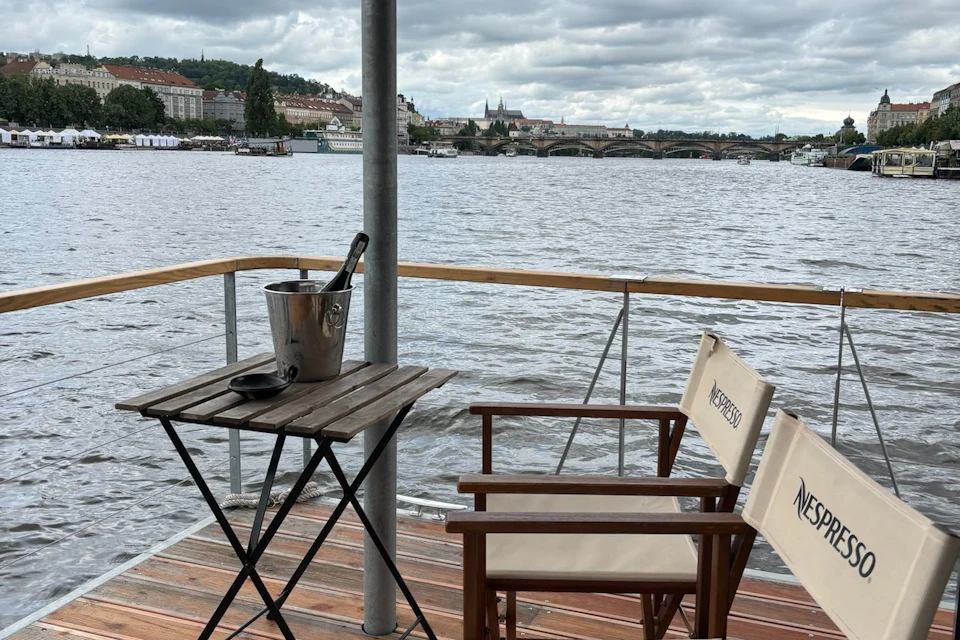 A wooden deck with a table holding an ice bucket and two chairs overlooking a river and a cityscape under a cloudy sky.