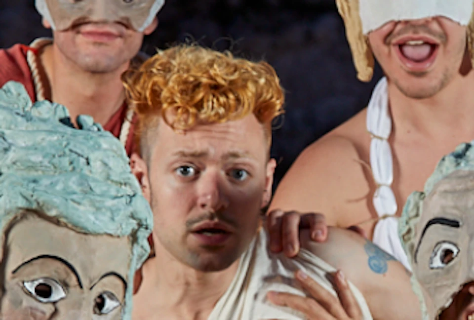 Five people in costume, wearing exaggerated masks and robes, performing with a historical building in the background.