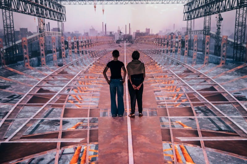 两个人站在一个生锈的大型建筑物上，后面有起重机和工业建筑。