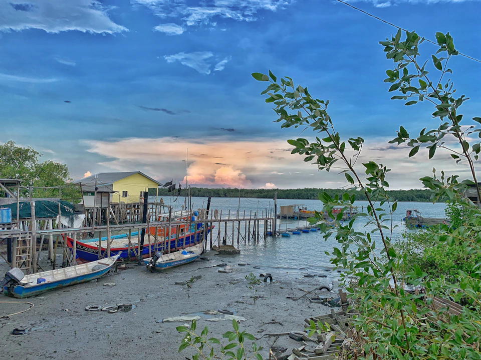 Colorful boats docked at a wooden pier, with a yellow house on stilts, under a blue sky with clouds. Greenery in the foreground.