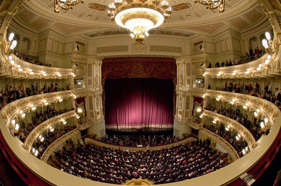A grand theater with a packed audience, ornate balconies, and a large chandelier, all facing a stage with closed red curtains.