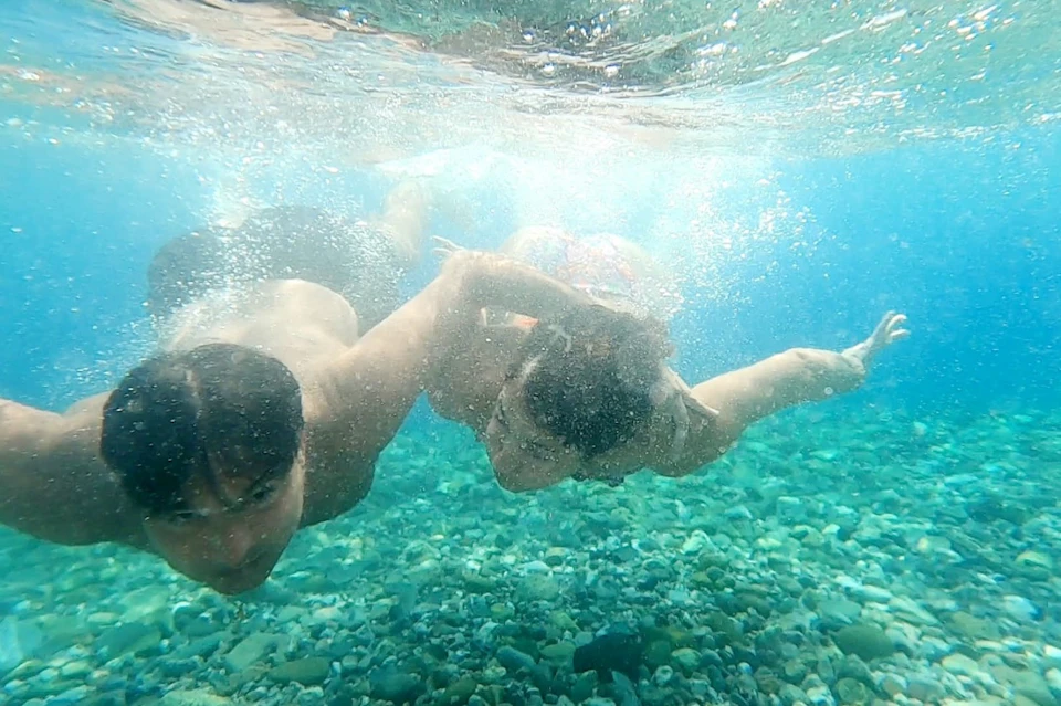 Two people swimming underwater above a rocky seabed in clear blue water, with bubbles surrounding them.