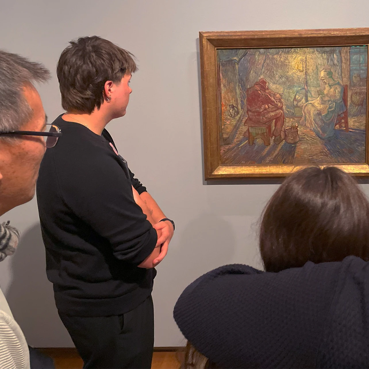 Three people observing a framed painting in a museum. The painting depicts a domestic scene with vibrant colors.