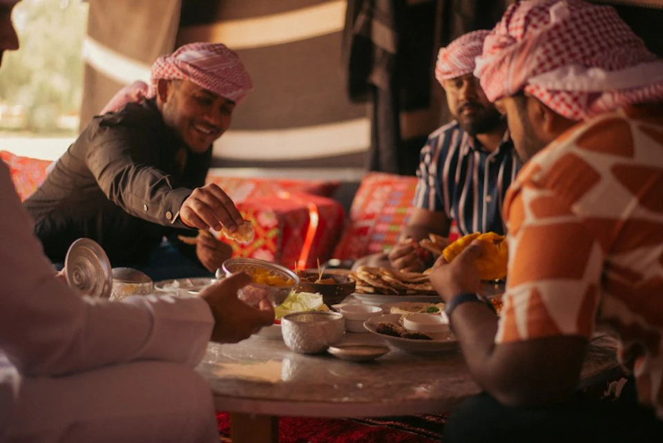 Four men in traditional clothing sit around a low table sharing a meal, with various dishes and flatbreads.