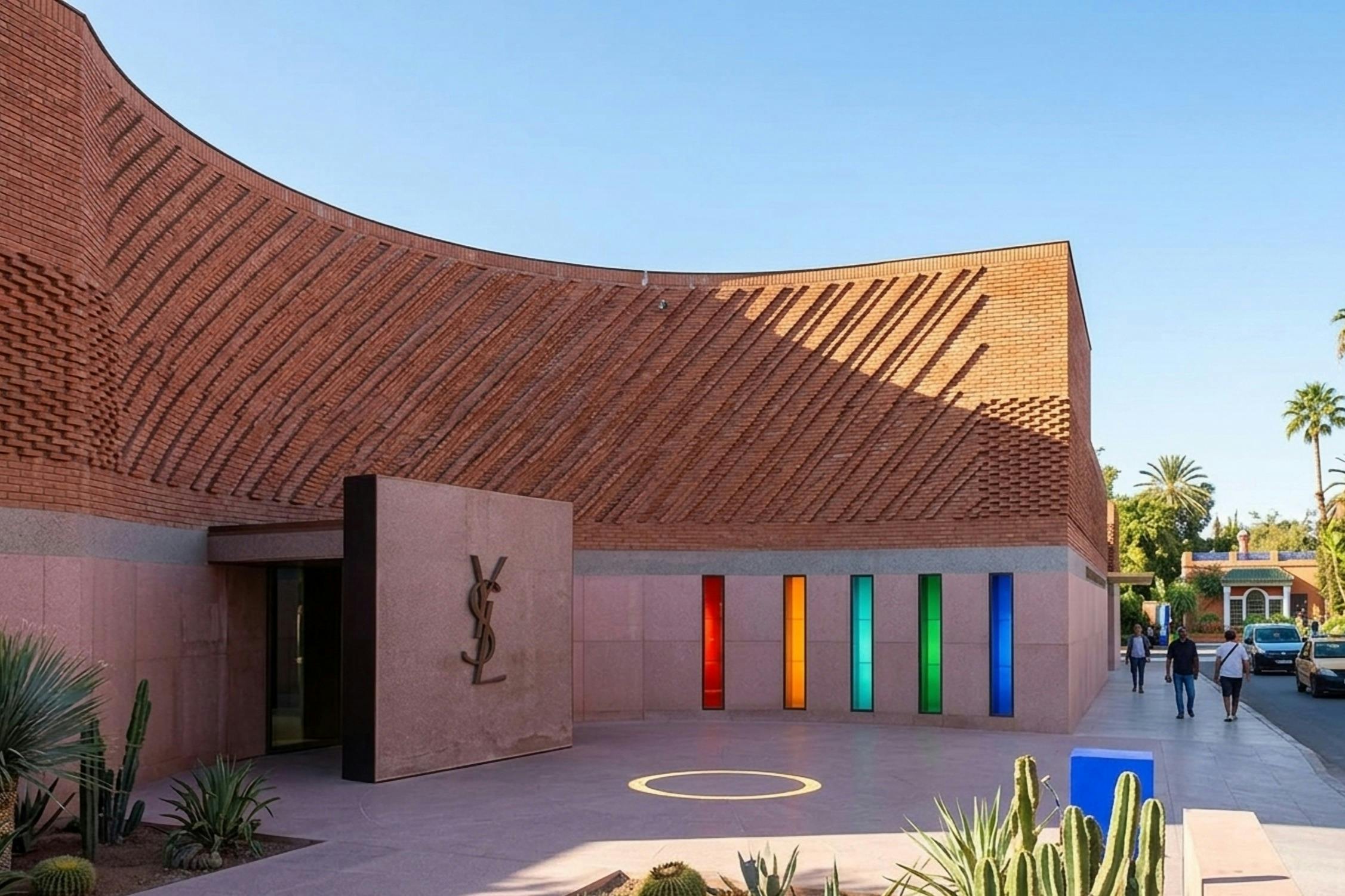 A modern brick building with colorful vertical glass panels, a YSL logo on a stone slab, and cacti in the foreground.