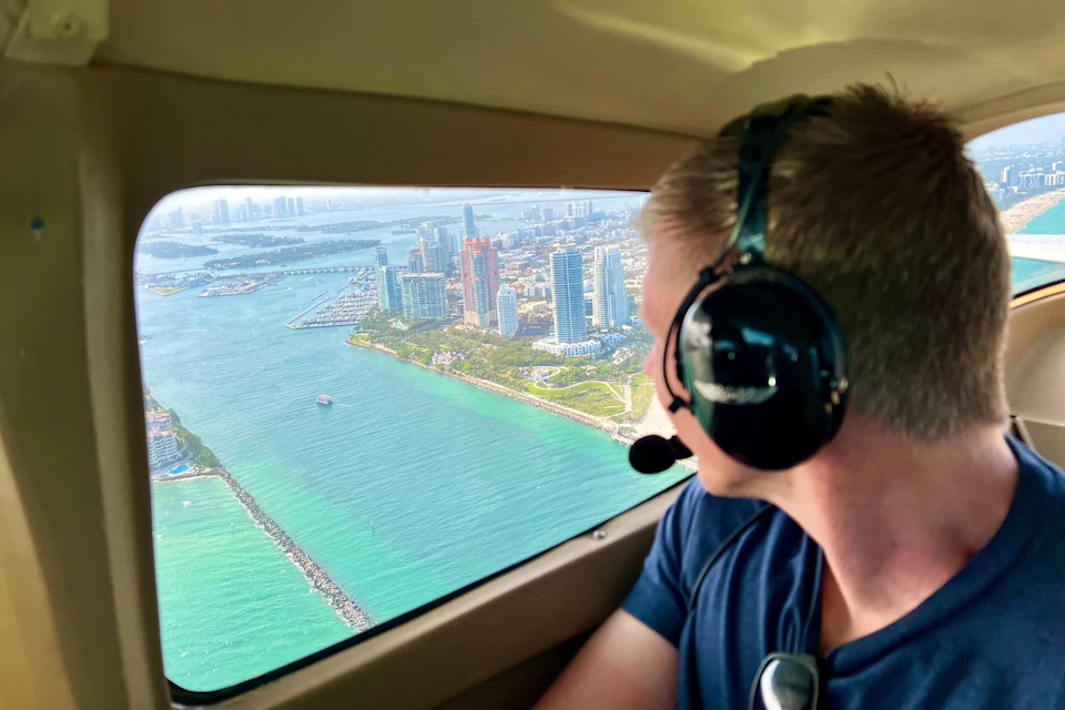 Person wearing a headset looks out of a small airplane window at a city with tall buildings and a coastline along turquoise waters.