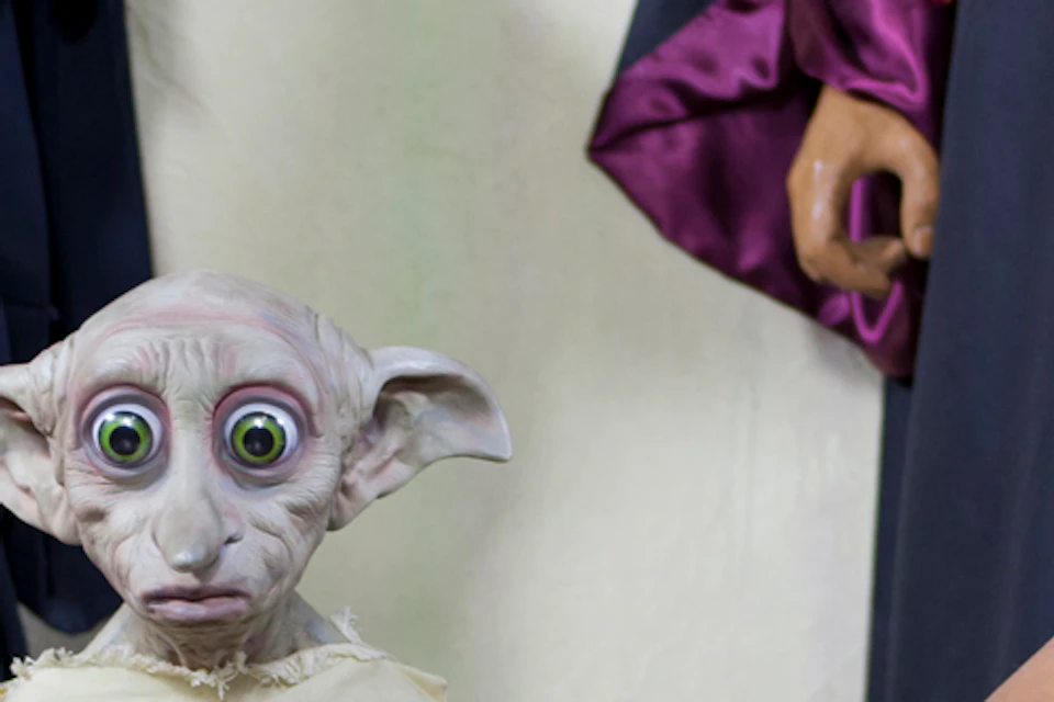 A girl kneels beside a Dobby statue from Harry Potter, striking a playful pose. Roped figures in costumes are in the background.