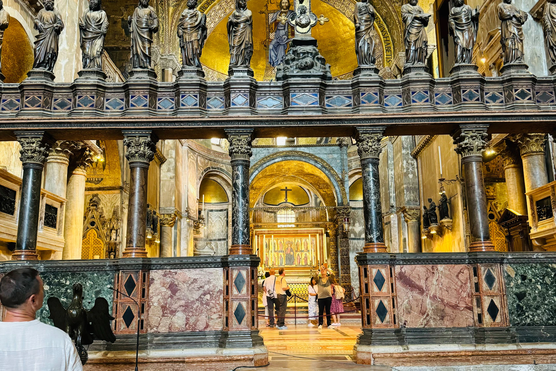 Interior of an ornate church with statues, arches, and people observing a golden altar under intricate decorations.