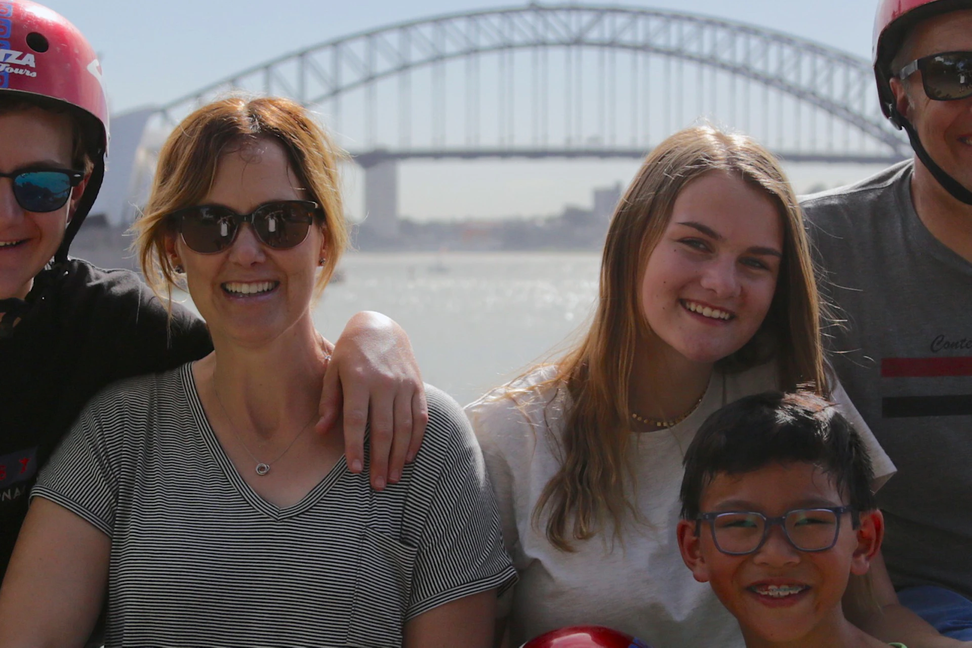 Group of five people smiling, posing near water with a bridge and cityscape in the background, some wearing helmets.