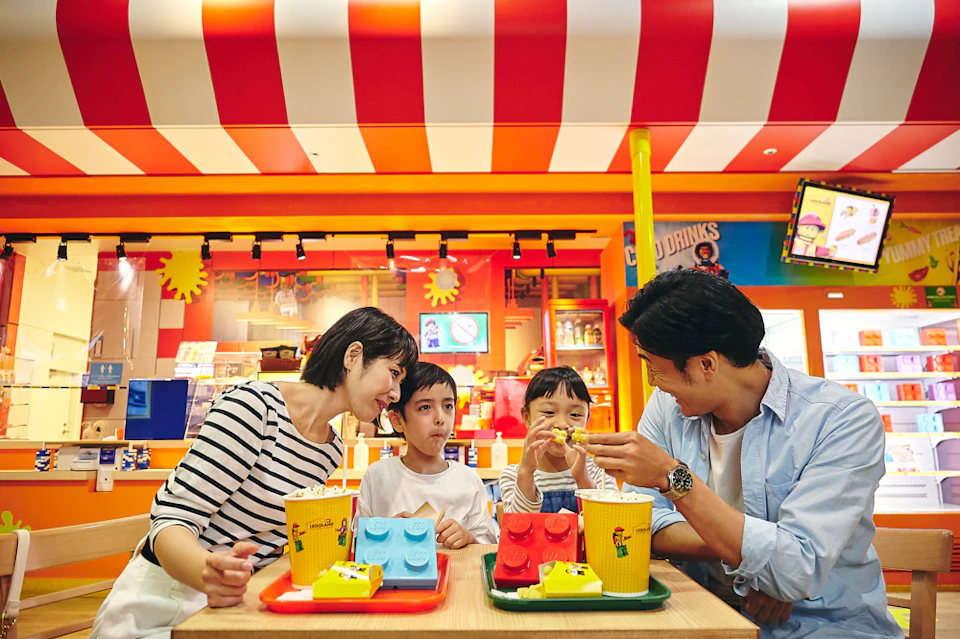 A family of four is eating at a vibrant LEGO-themed restaurant with red and white striped decor. They have LEGO-shaped dishes.