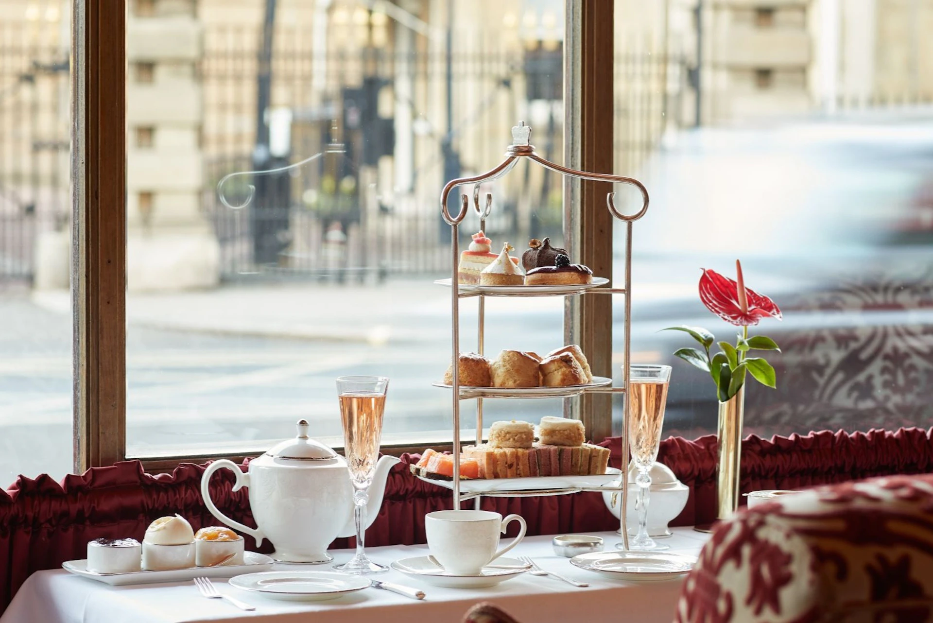 A table set with tea, pastries on a tiered stand, and champagne glasses by a window overlooking a street scene.