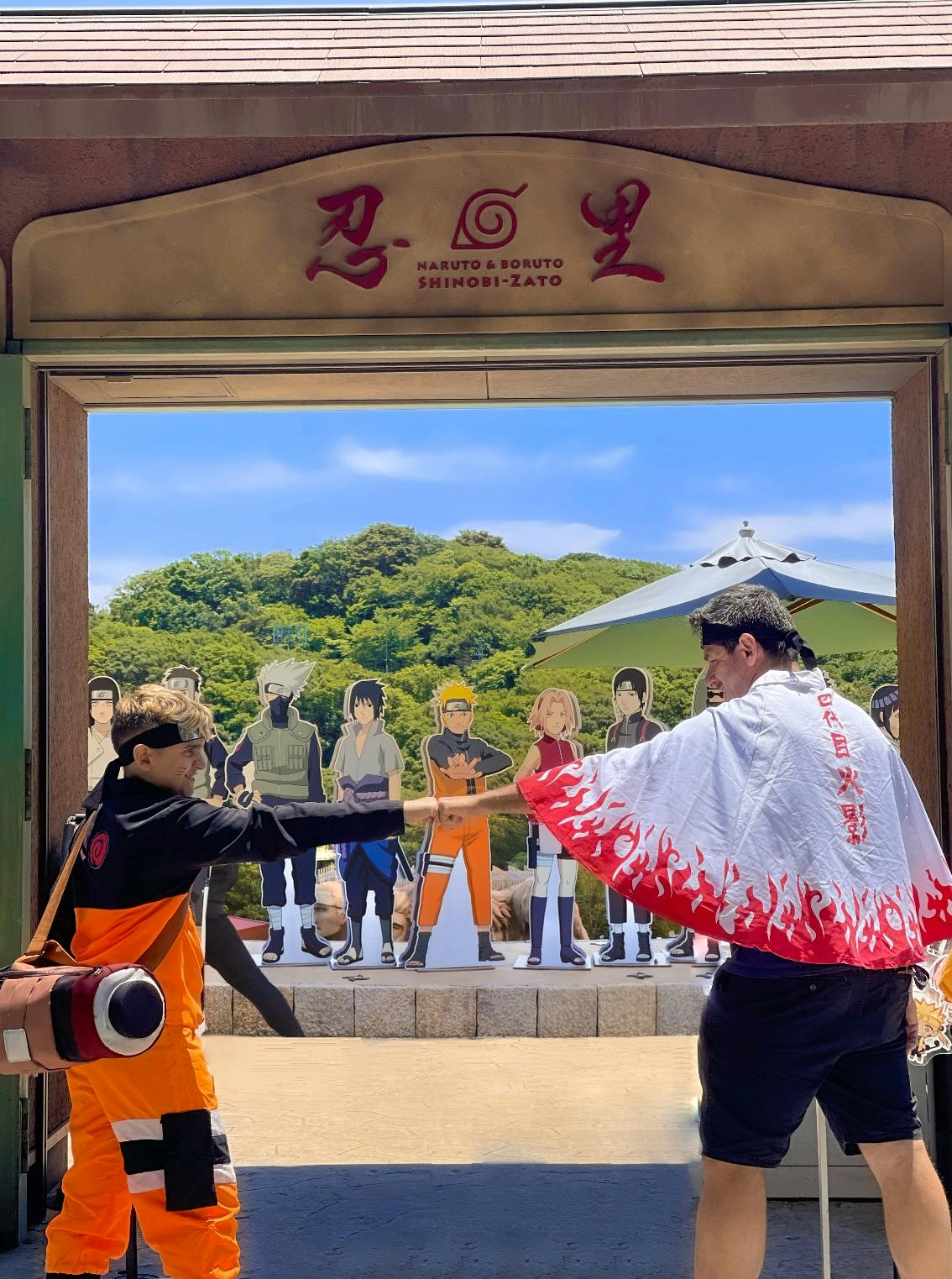 Two people in cosplay costumes pose in an outdoor area with Naruto-themed decorations and cutouts of characters in the background.