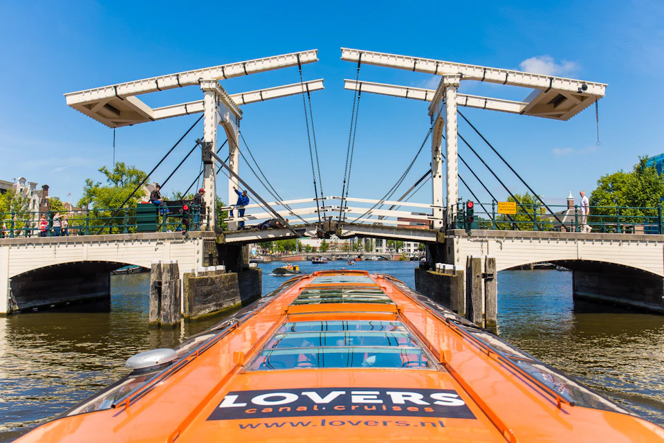Bridge opening over a canal as an orange Lovers Canal Cruise boat approaches. People stand on the bridge under a clear blue sky.