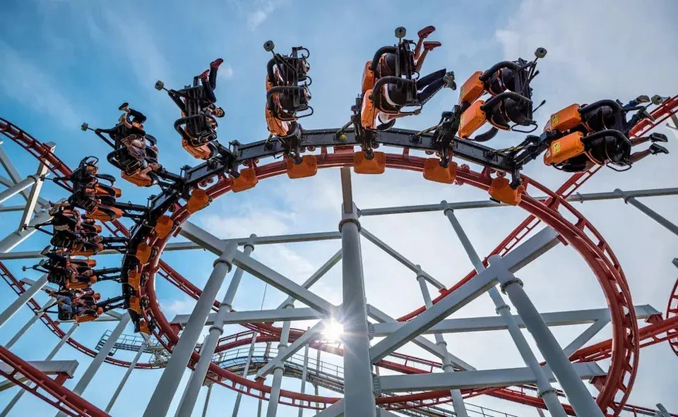 People riding an inverted roller coaster with red and orange tracks against a clear blue sky.