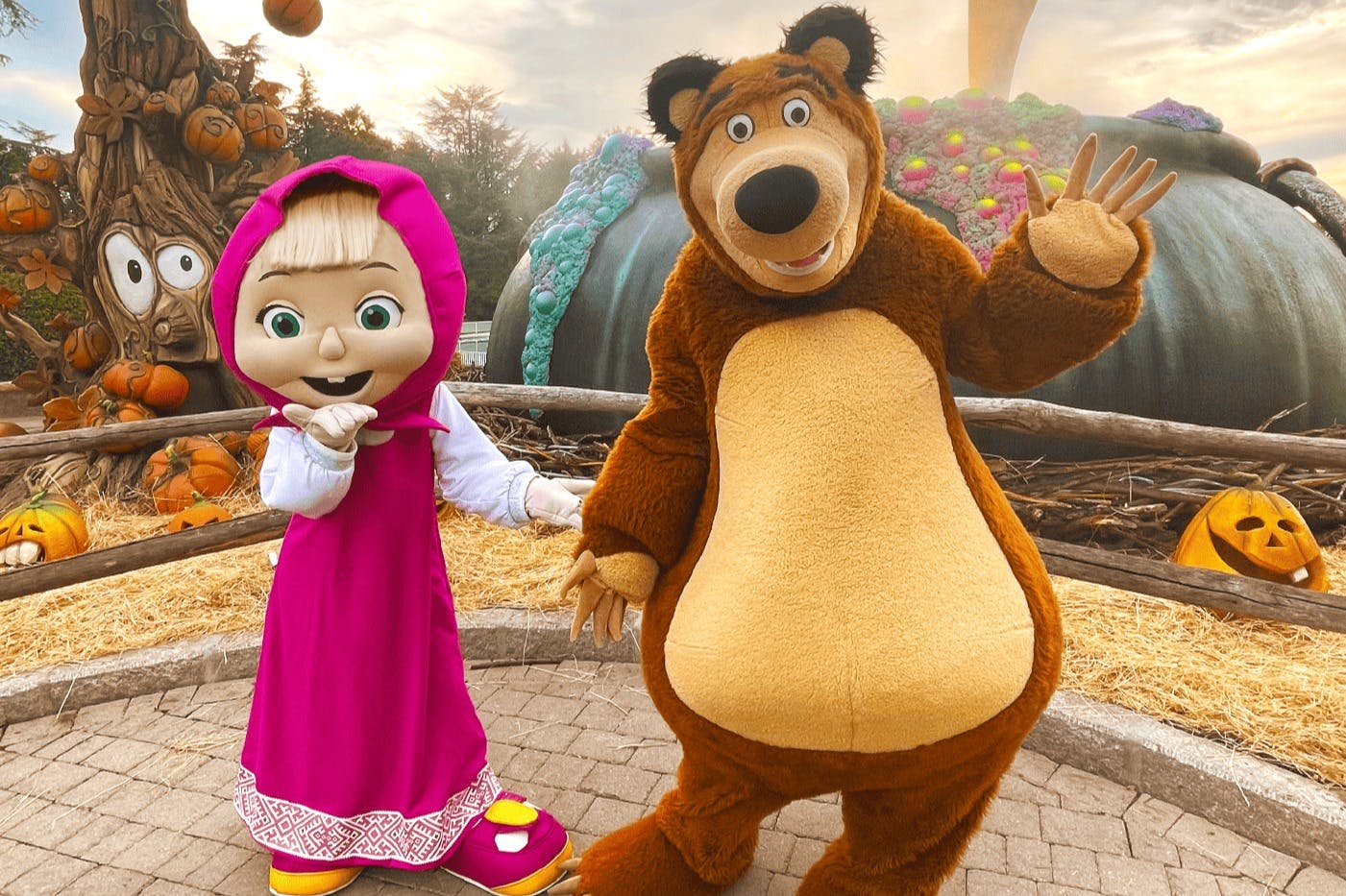 Δύο μεταμφιεσμένοι χαρακτήρες, ένα κορίτσι με ροζ φόρεμα και μια αρκούδα, ποζάρουν σε ένα διακοσμημένο, φθινοπωρινό σκηνικό.