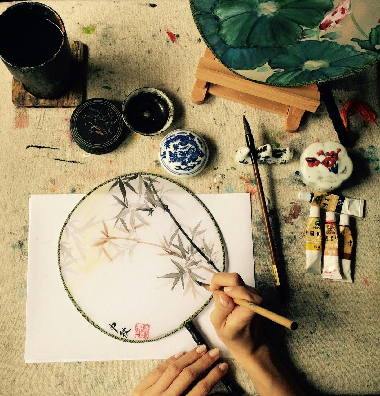 丸い扇の上に竹を描く手。 近くには、塗装用チューブ、ブラシ、インクポット、花柄の扇風機が完備されています。