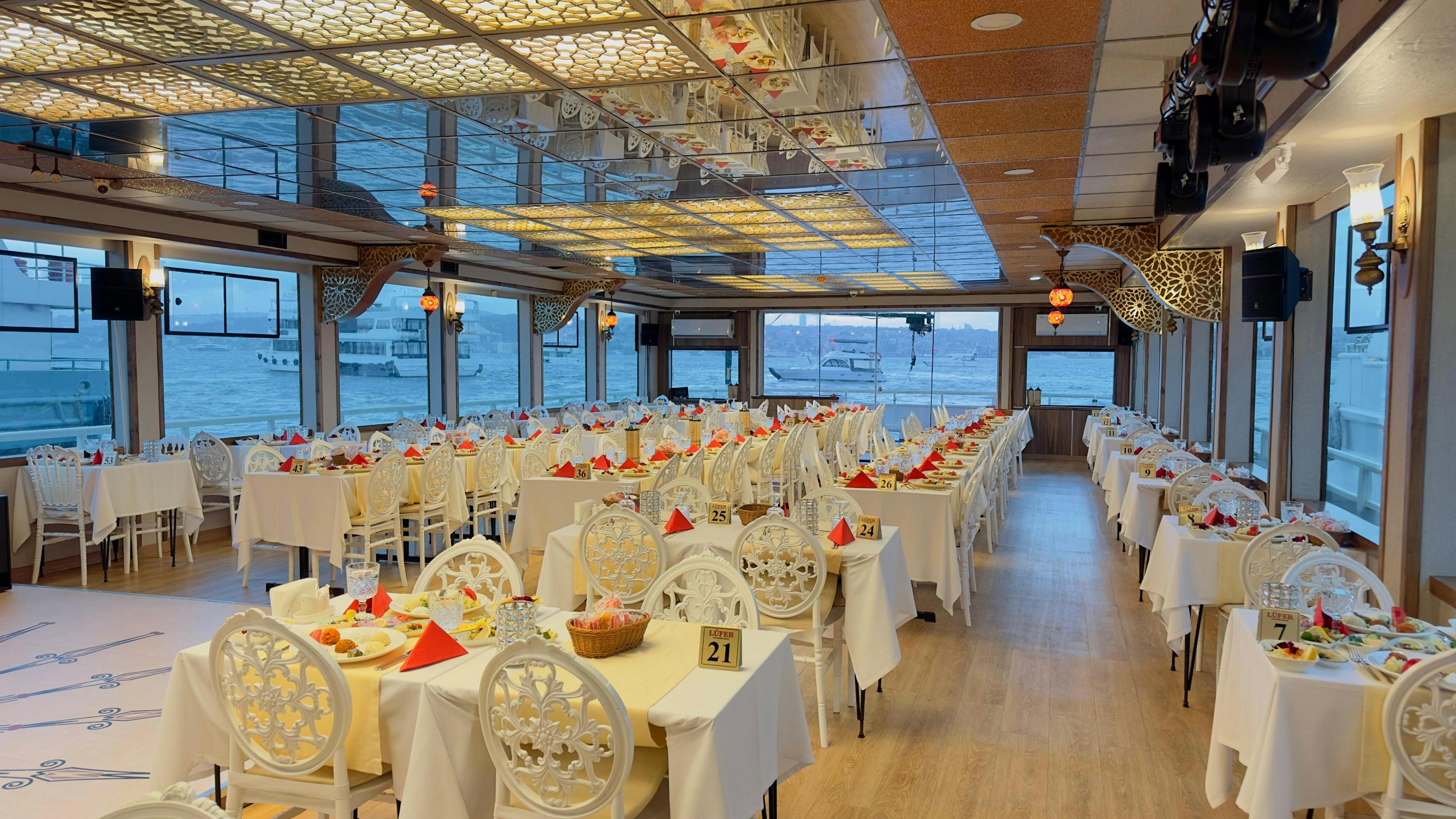 A spacious restaurant interior with white tables and chairs, set with red napkins, overlooking a water view through large windows.