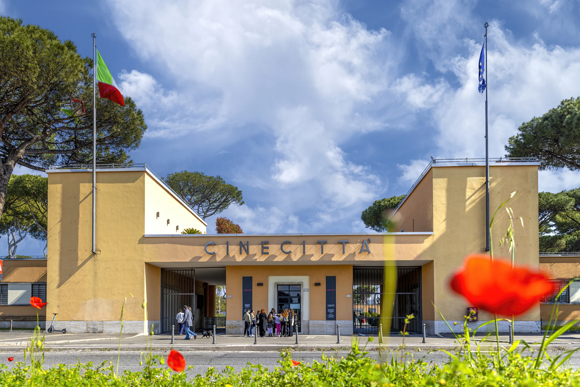 Entrance to the Cinecittà studios with a group of people outside, set against a bright sky with flags and trees visible.
