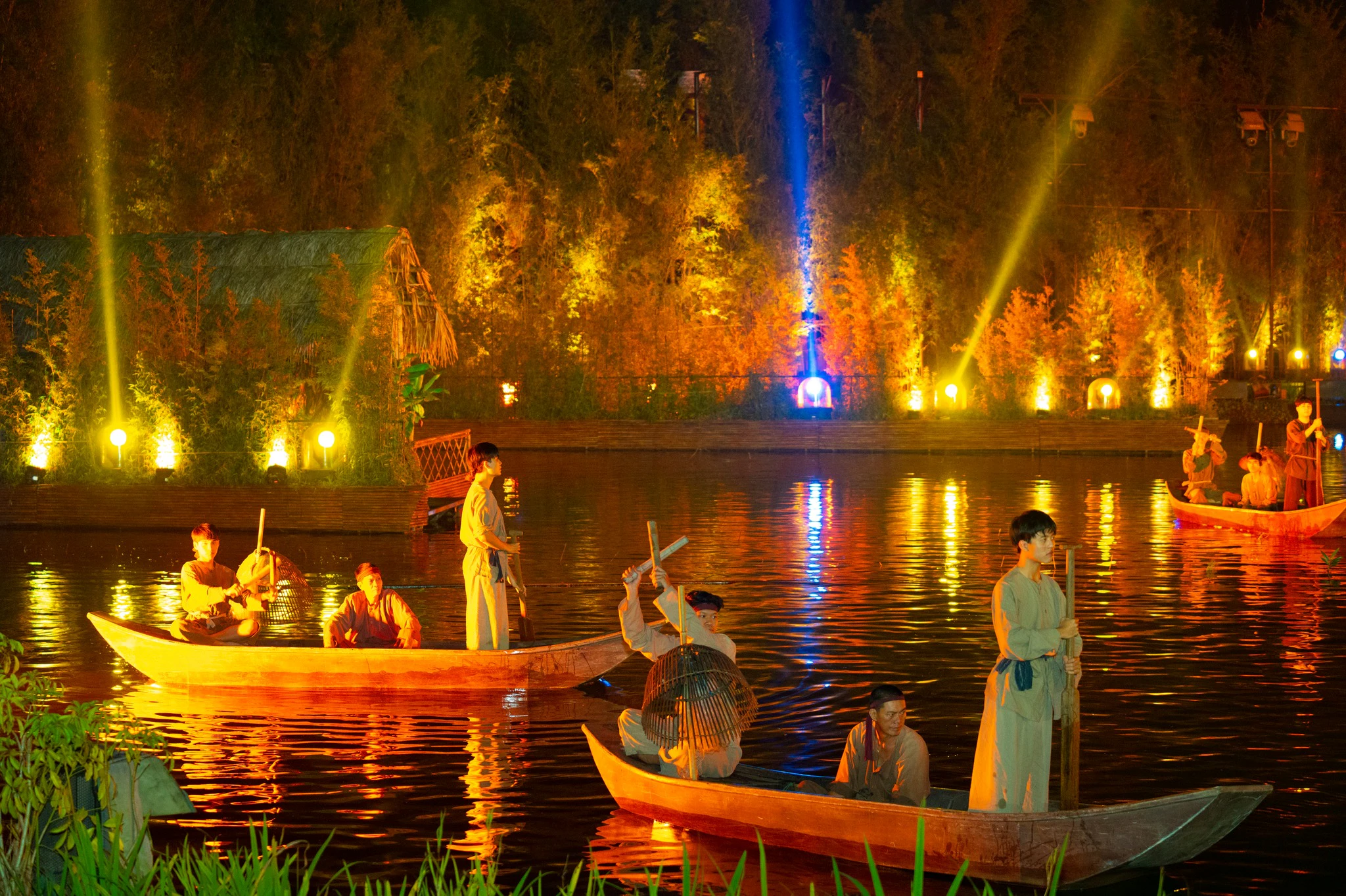 People in traditional clothing on wooden boats in a brightly lit river scene with trees and illuminated structures.