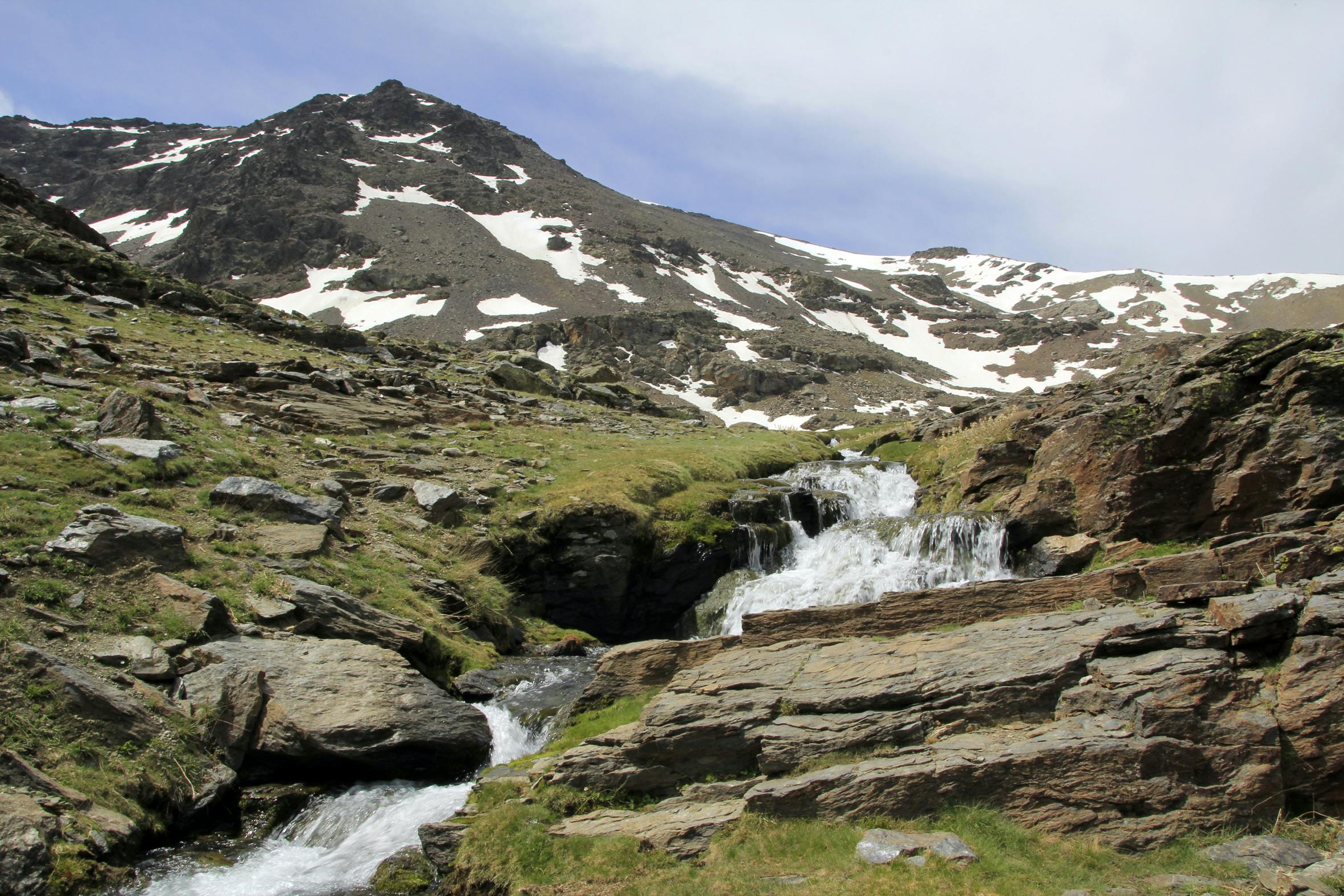 Mountain landscape with patches of snow, a rocky terrain, and a small waterfall flowing down grassy slopes under a partly cloudy sky.