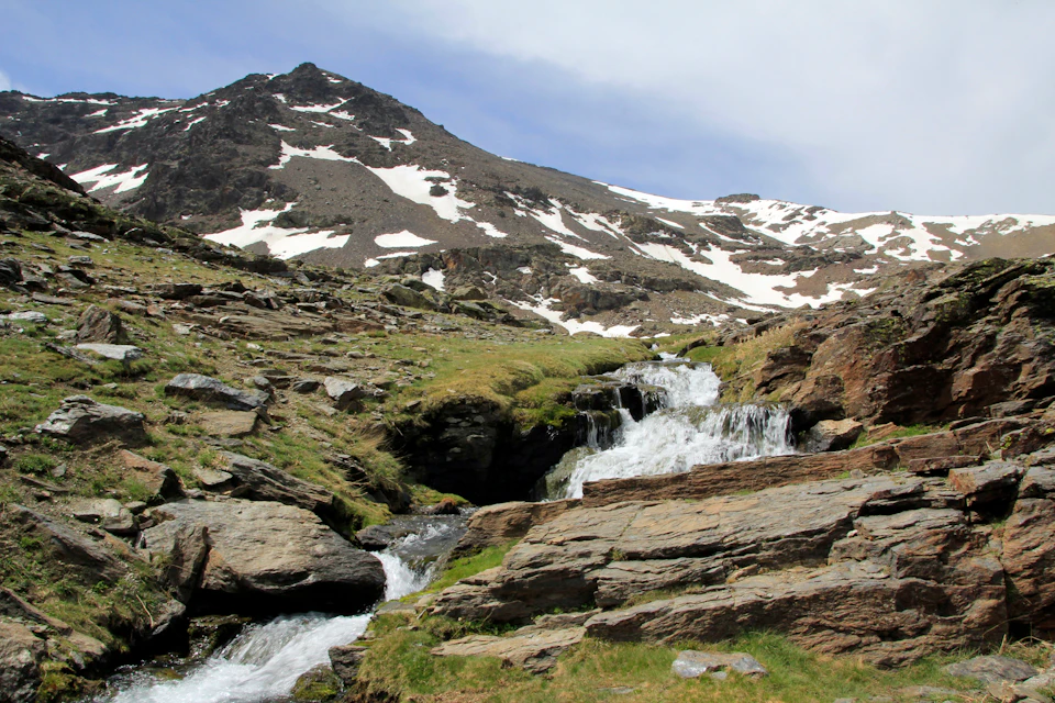 Mountain landscape with patches of snow, a rocky terrain, and a small waterfall flowing down grassy slopes under a partly cloudy sky.