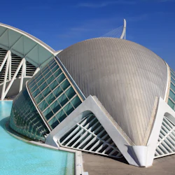 En modern, futuristisk arkitektonisk struktur med böjda glasväggar och en reflekterande pool under en klarblå himmel.