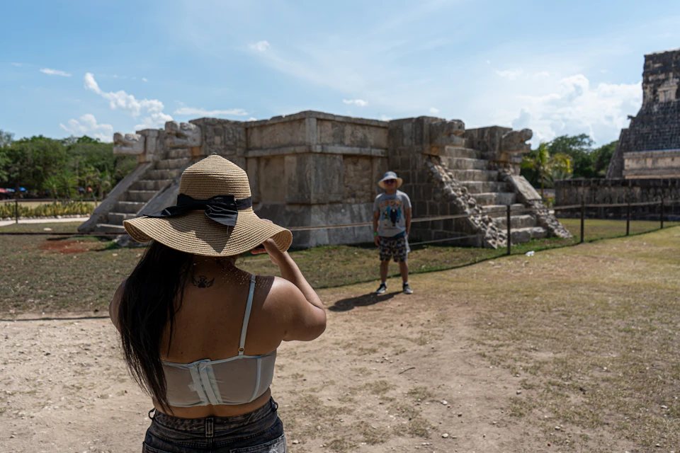 A person in a hat takes a photo of another in front of a stone temple with stairs against a clear blue sky.