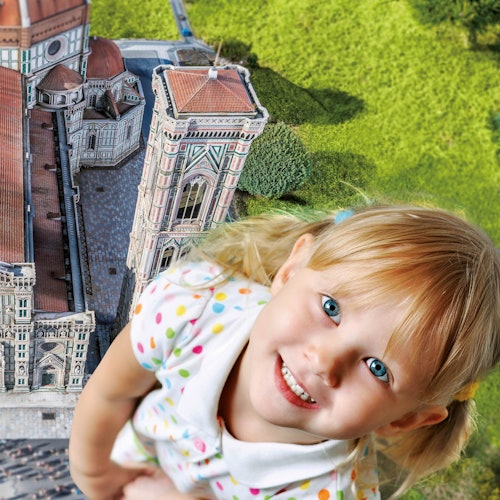Una giovane ragazza sorridente dai capelli biondi appare gigante accanto a un dettagliato modello in miniatura di un edificio storico e a una vegetazione lussureggiante.