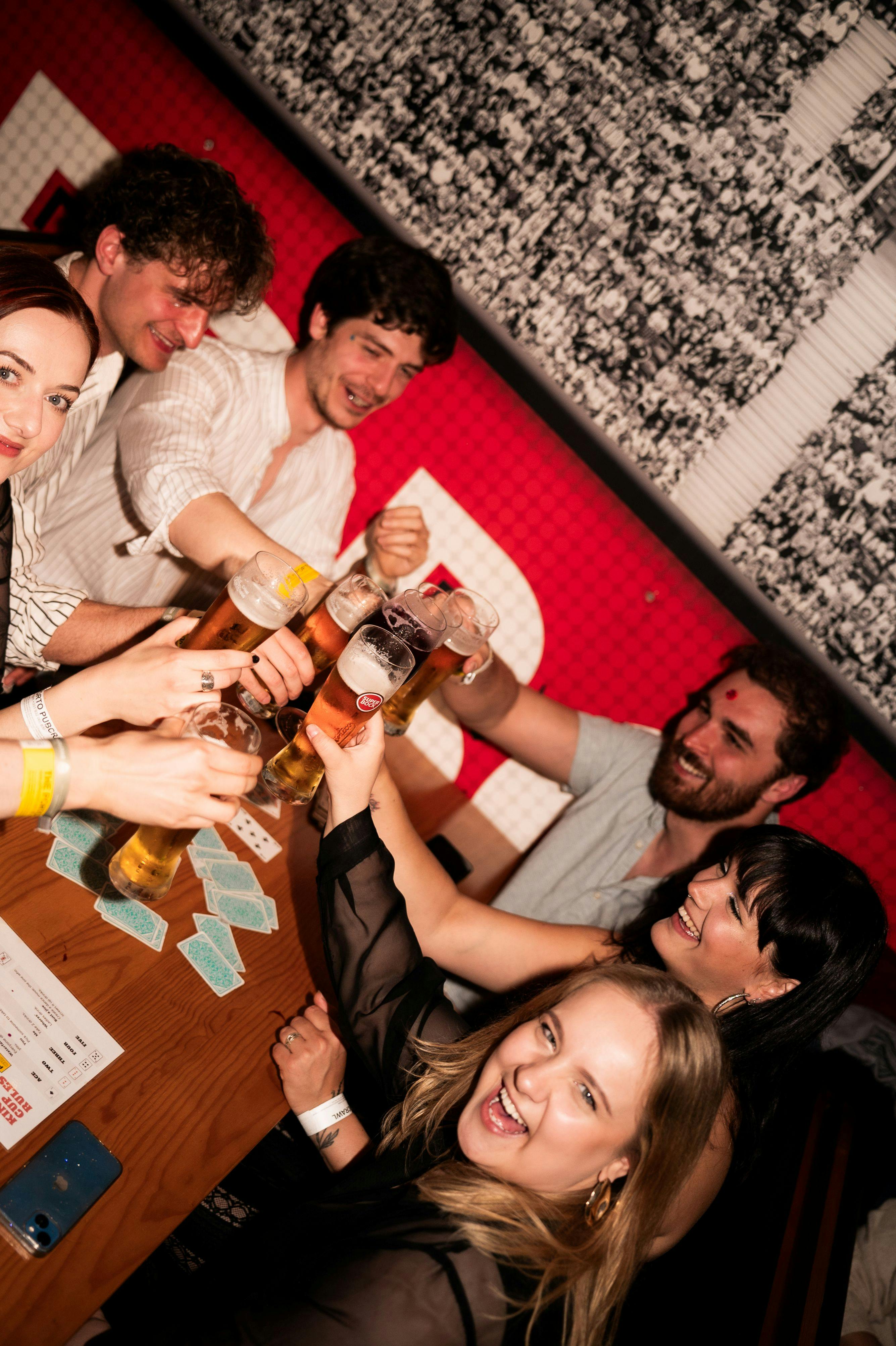 Een groep mensen rond een tafel die een glas bier heffen in een feestelijke toost, met een rode muur op de achtergrond.