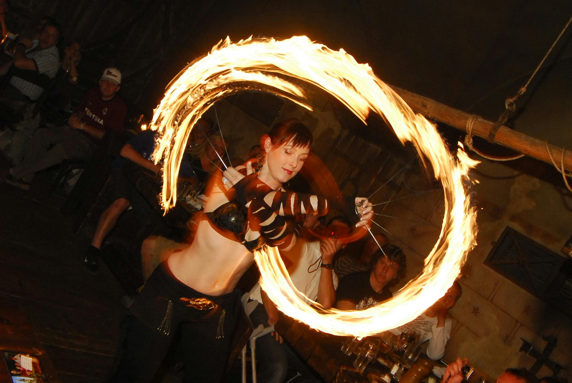A performer swings fiery objects in circular patterns, entertaining seated audience members in a dimly-lit venue.