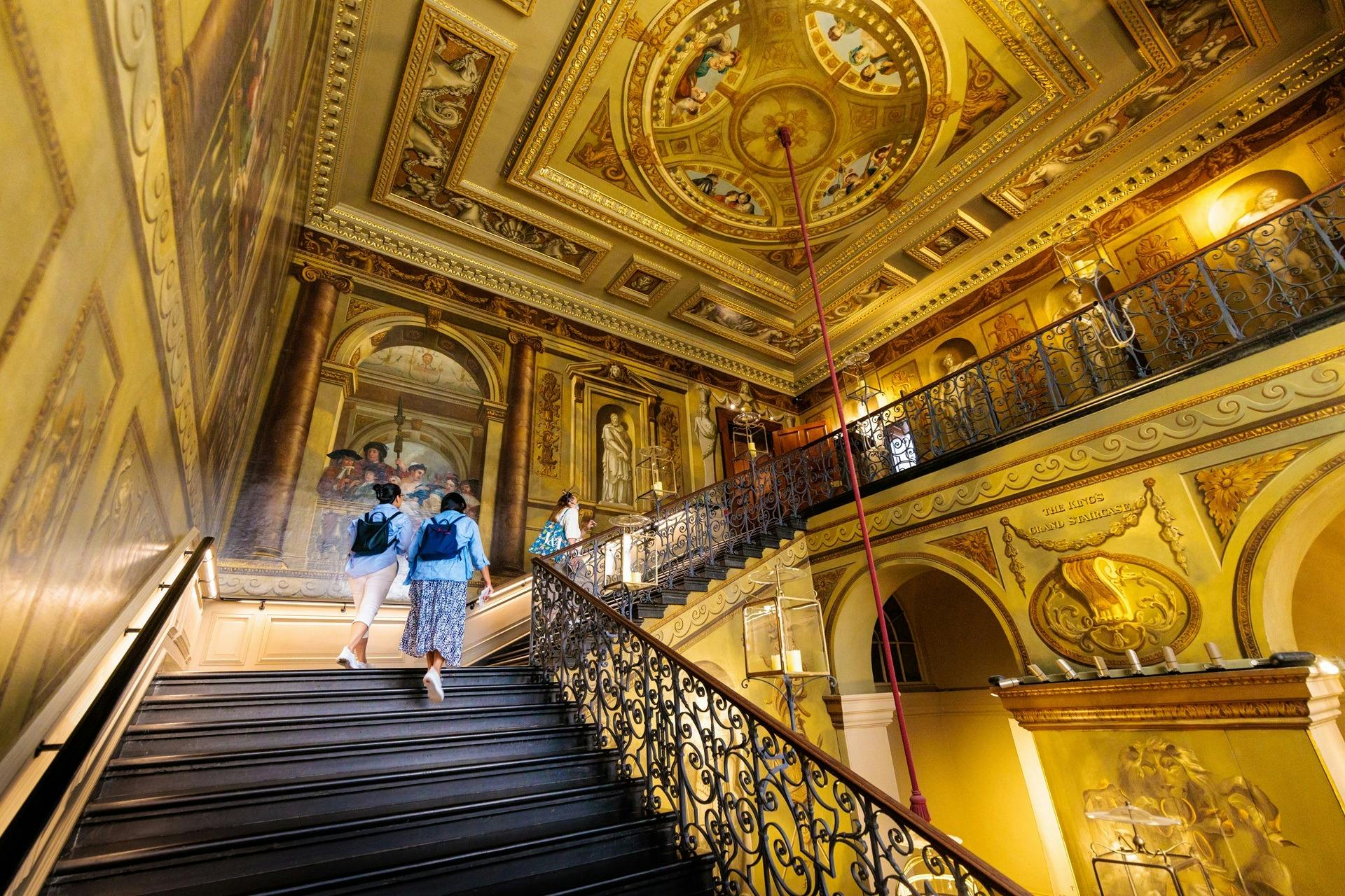 Two people with backpacks ascend an ornate staircase in a grand hall adorned with elaborate murals and detailed ceiling artwork.