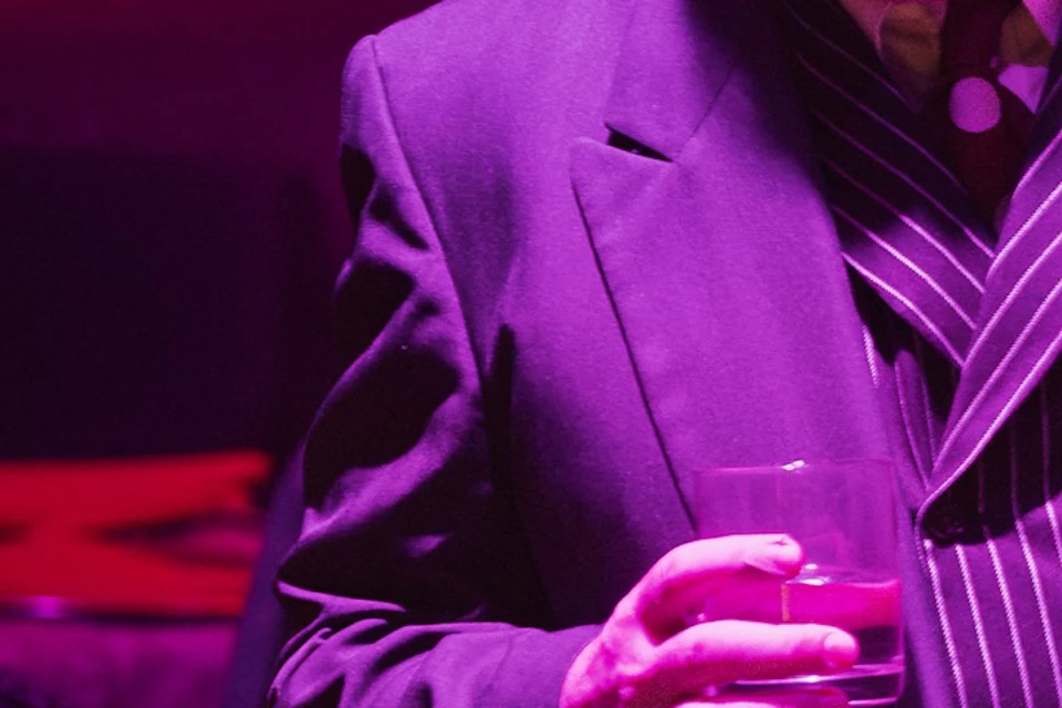 A person in a formal suit holding a drink, standing in a dimly lit setting with red curtains and a lamp nearby.