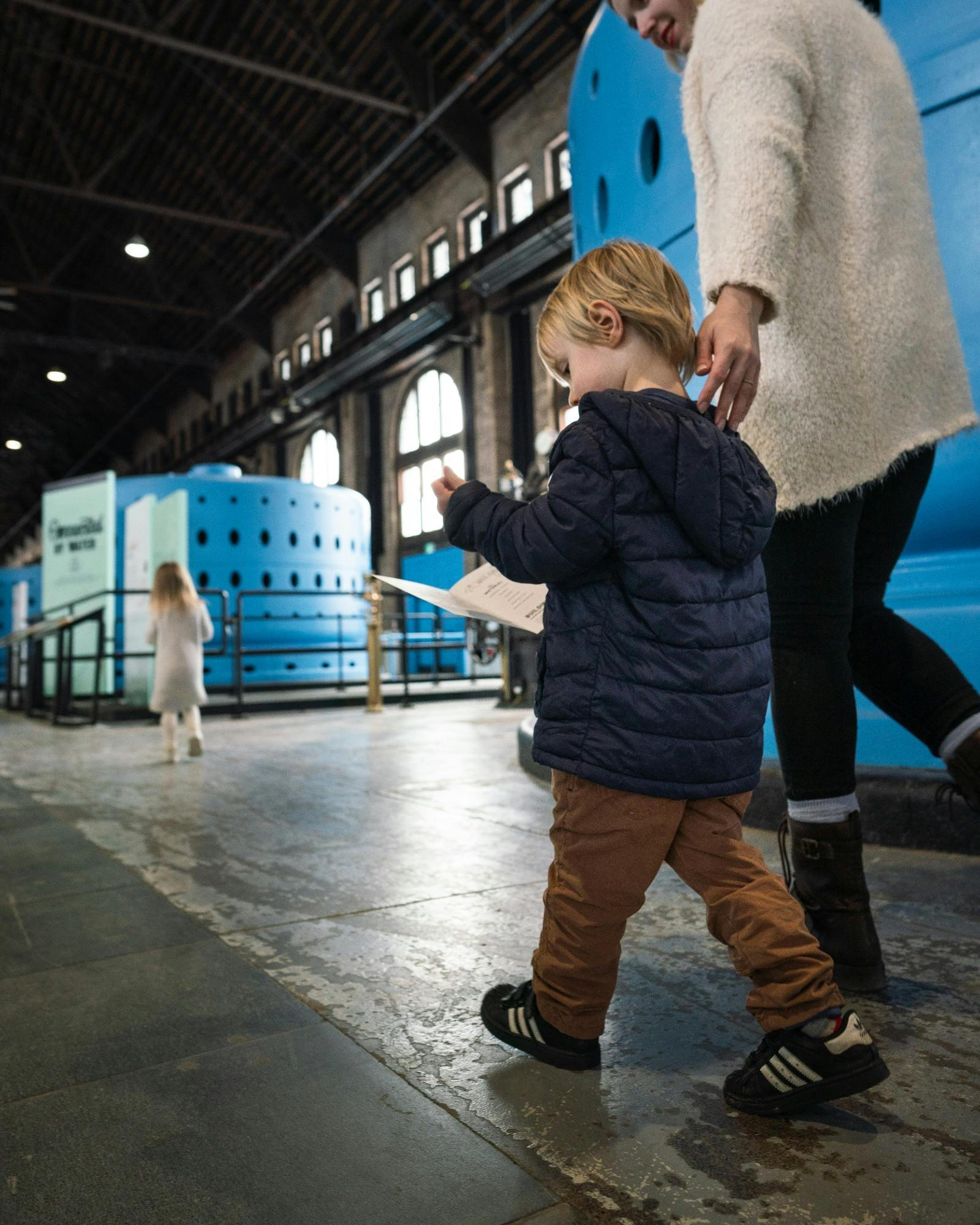 Et barn i en blå jakke går indendørs ved siden af en kvinde, med stort blåt industriudstyr og en anden person synlig i baggrunden.